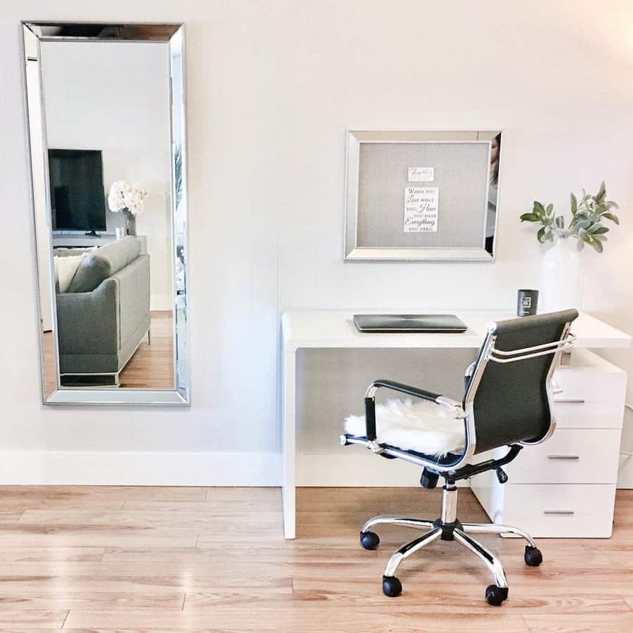 Minimalist workspace with a white desk, black chair, laptop, framed quotes, and a tall mirror reflecting a living room