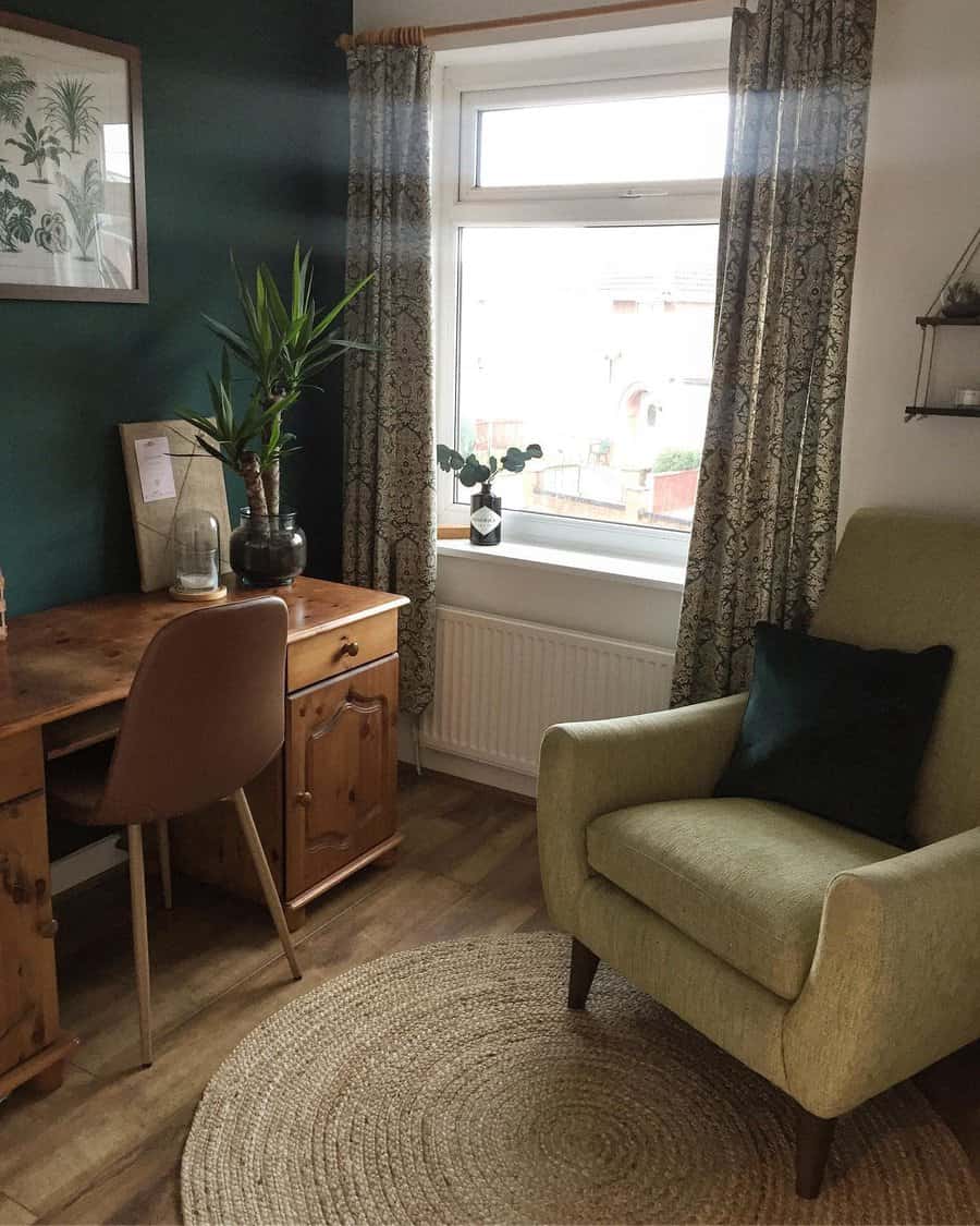 Cozy room with a wooden desk and brown chair, green armchair, potted plants, round rug, and a window with patterned curtains