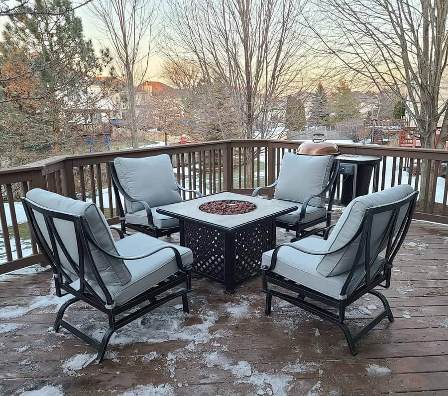 Outdoor deck with a fire pit table surrounded by four cushioned chairs, light dusting of snow, trees and houses in the background