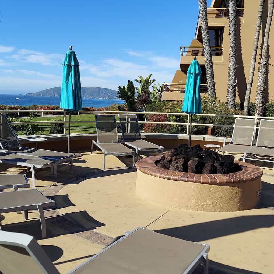 Outdoor patio with lounge chairs and a round fire pit. Blue parasols are closed. Ocean and hills visible in the background