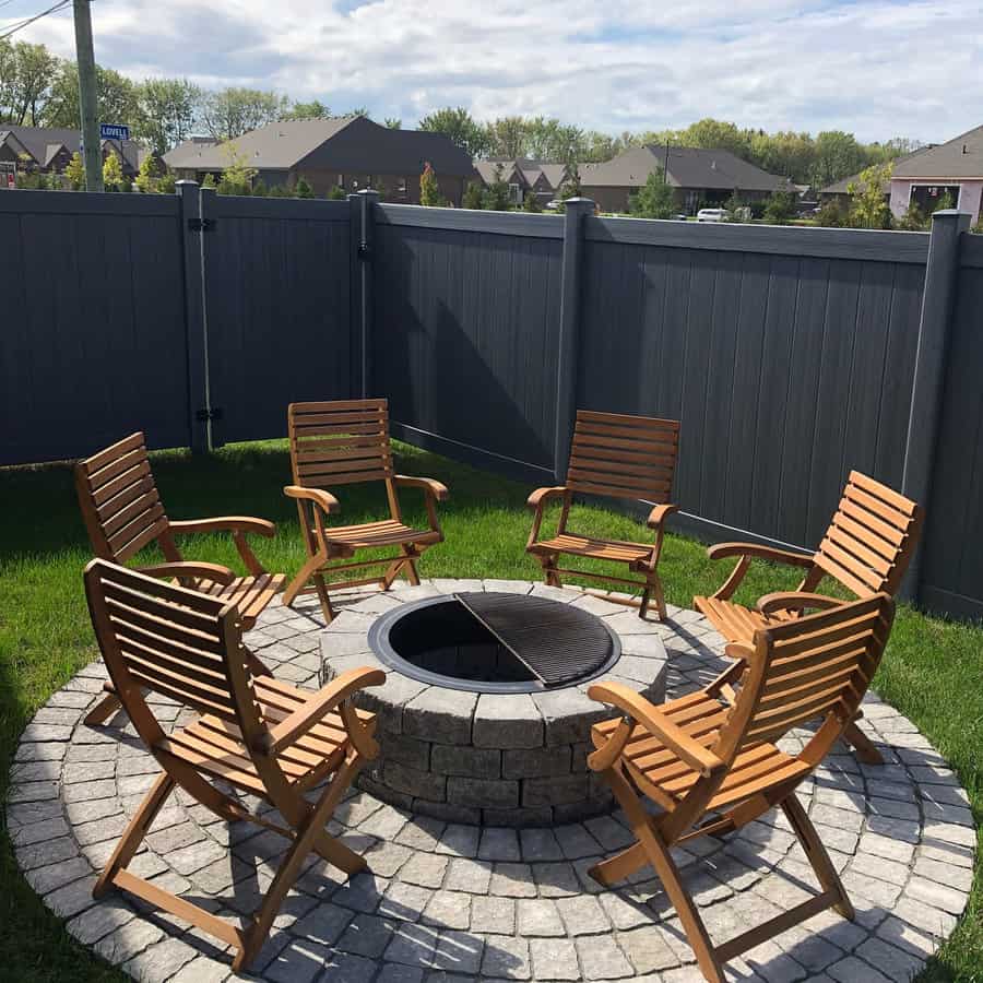 Wooden chairs arranged around a stone fire pit on a circular patio in a backyard with a tall fence and houses in the background