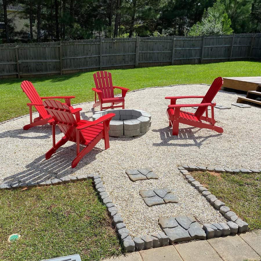 Four red Adirondack chairs surround a stone fire pit on a gravel patio, with a wooden fence and trees in the background