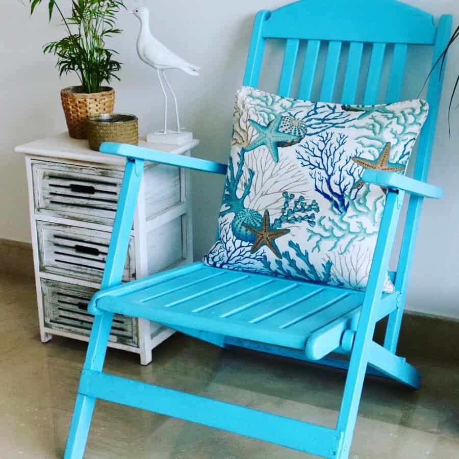 Bright turquoise wooden chair with a coastal-themed cushion, styled next to a distressed white side table with woven decor and greenery.