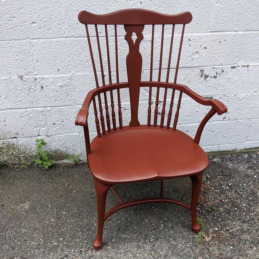 Classic Windsor-style chair painted in deep terracotta, featuring elegant spindle details and curved armrests, set against a textured wall.