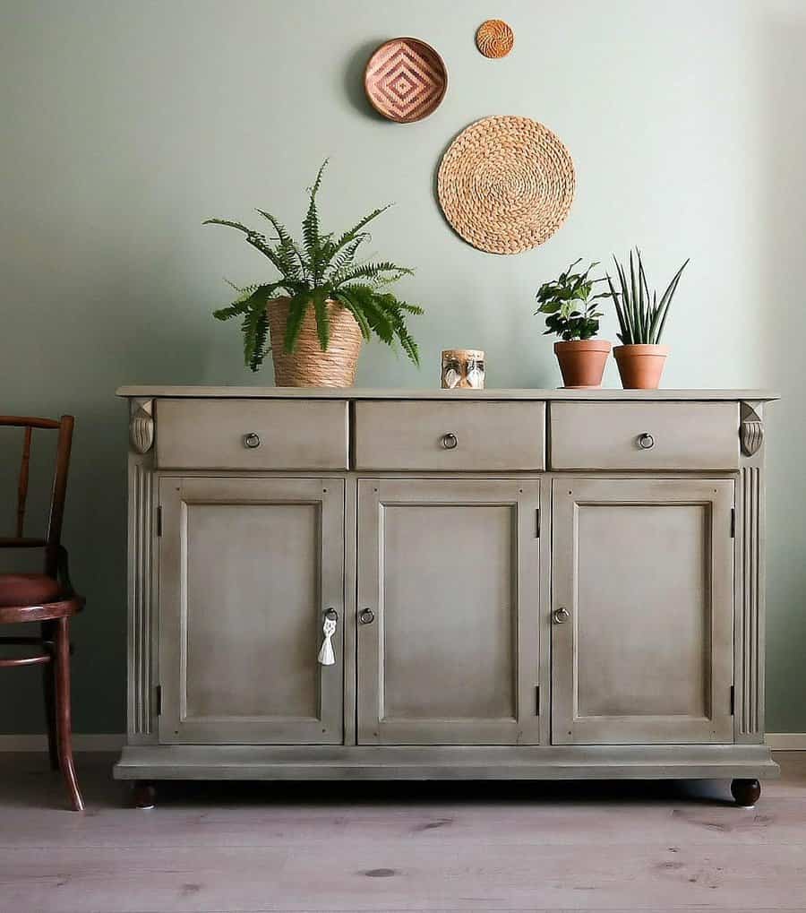 Vintage sideboard painted in muted sage green with a distressed finish, styled with woven wall decor, potted plants, and earthy accents.