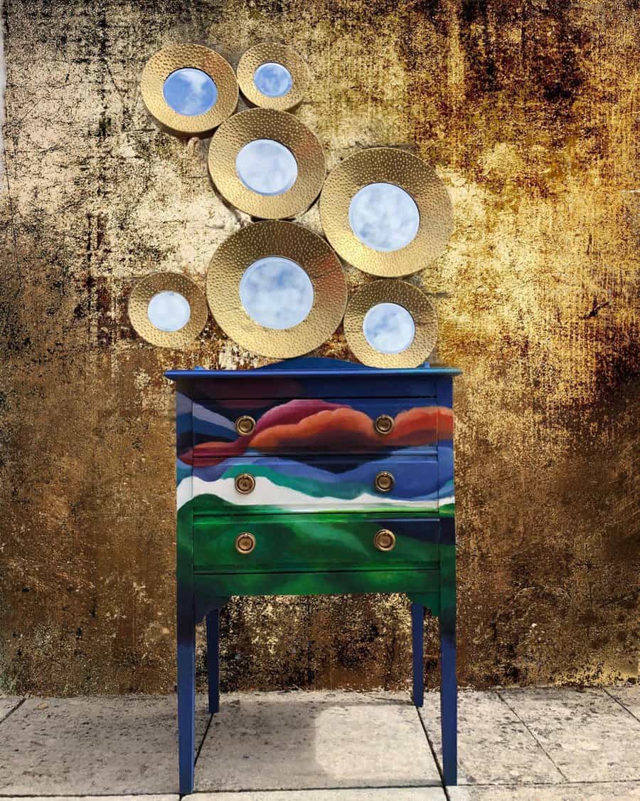 Bold hand-painted dresser with a colorful abstract landscape design, paired with gold circular mirrors against a textured metallic wall.