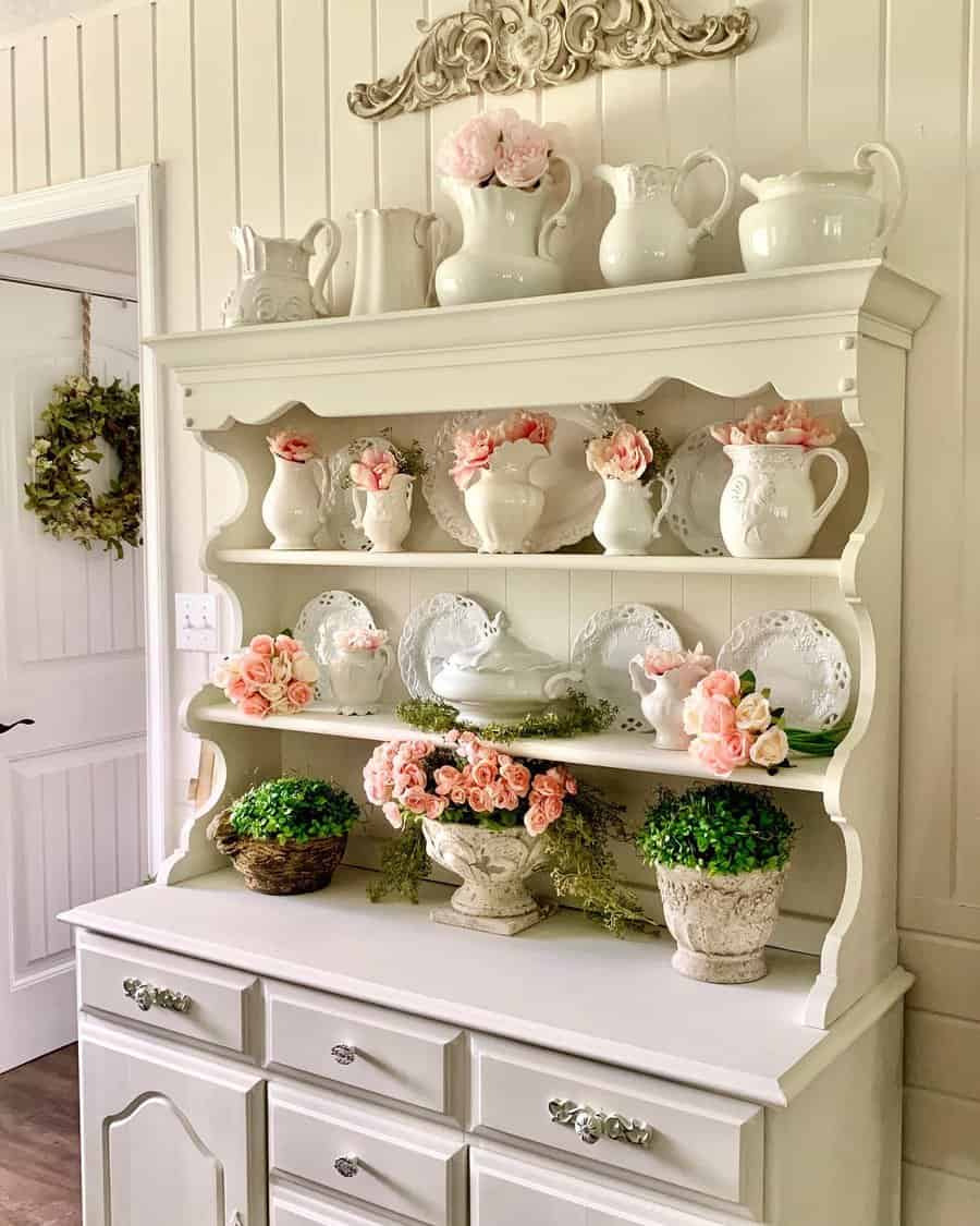 Elegant cream-painted hutch adorned with vintage white pitchers, lace-edged plates, and pink floral arrangements in a French farmhouse setting.