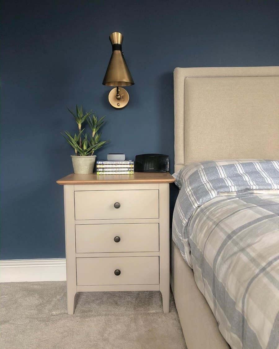 Modern bedside table painted in soft beige with a wooden top, styled with a potted plant and books, set against a navy blue wall with a brass sconce.