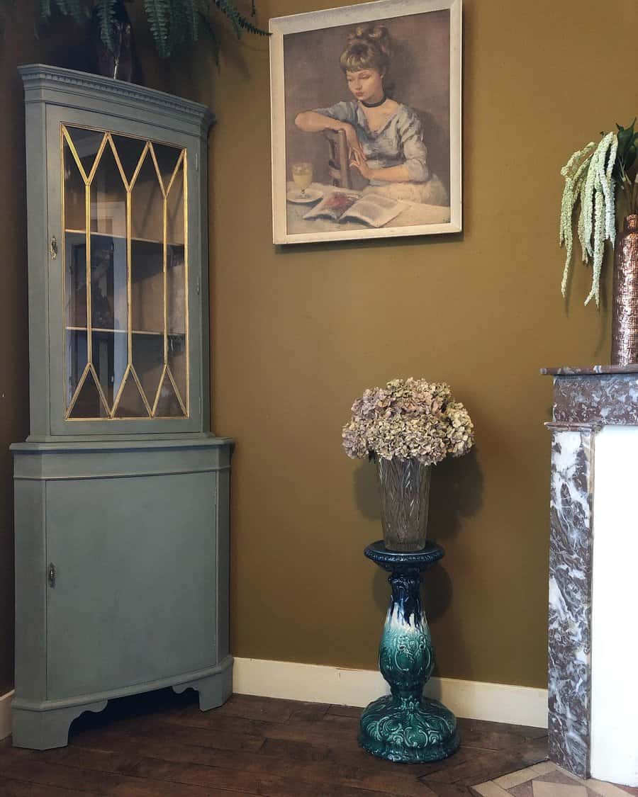 Vintage corner cabinet painted in muted sage green with gold detailing, styled in a classic room with an ornate pedestal and framed artwork.