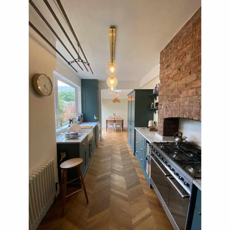 Galley kitchen with wooden herringbone floor, exposed brick wall, teal cabinets, large window, and a dining area at the end