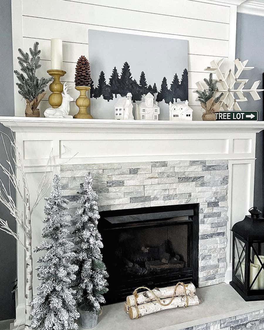 Mantel with winter decor: pine trees artwork, mini white houses, candles, pinecones, snowflakes, small flocked trees, and logs