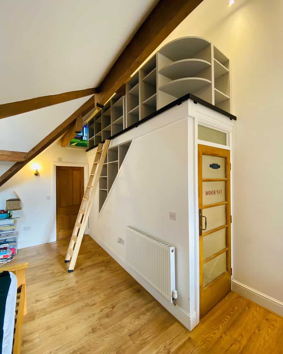 Loft-style attic storage with built-in open shelves above a small room, accessed by a wooden ladder, maximizing vertical space efficiently.