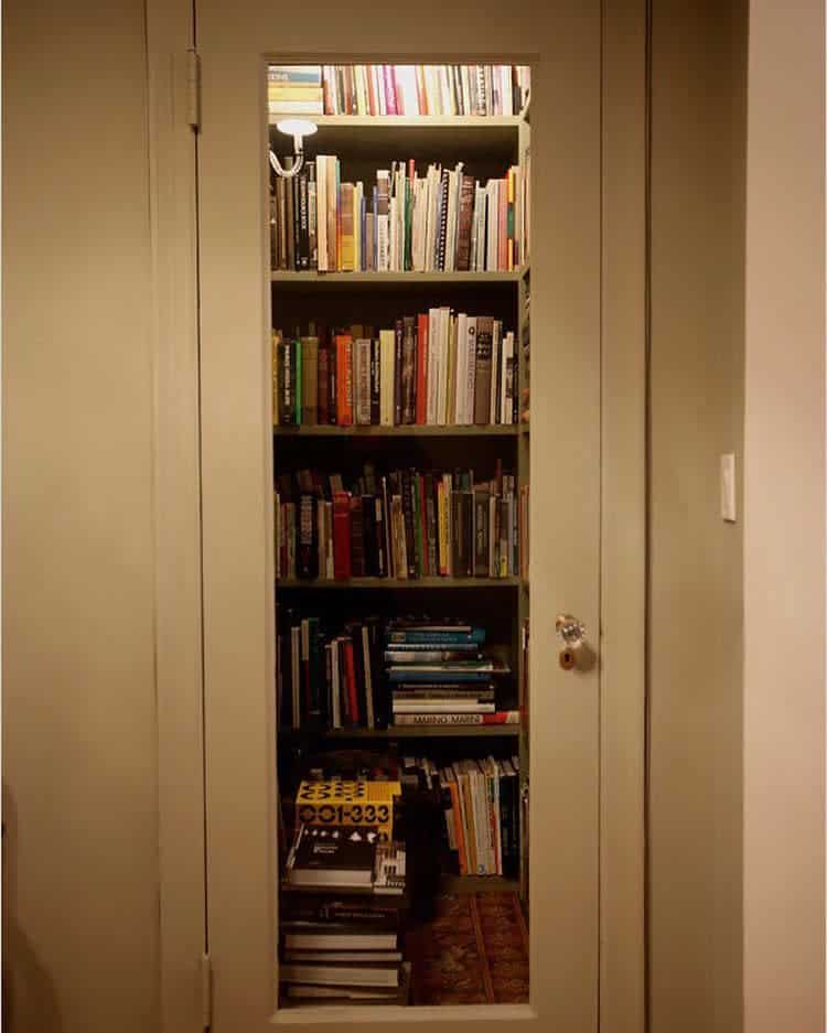 A hidden book closet with neatly arranged books on multiple shelves, illuminated by a small lamp, behind a door with a glass panel.