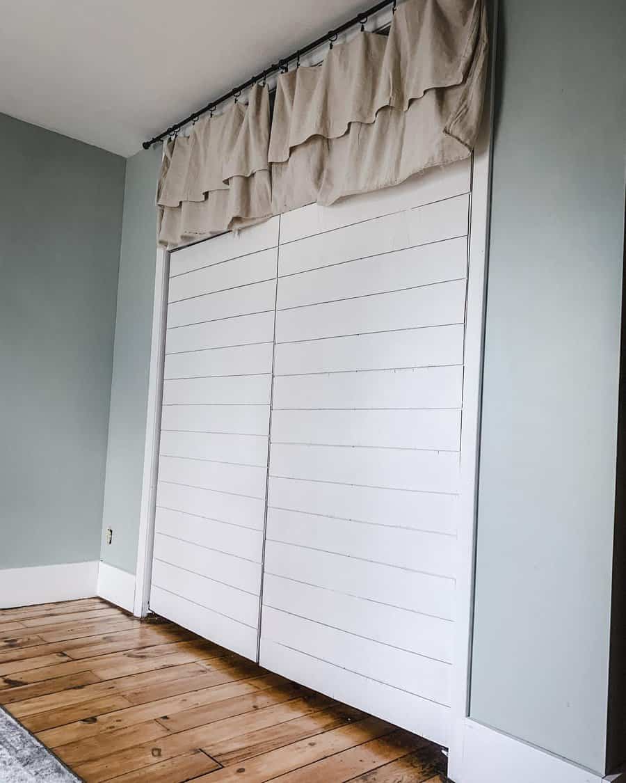 White shiplap doors with a beige valance at the top in a room with light blue walls and wooden floor