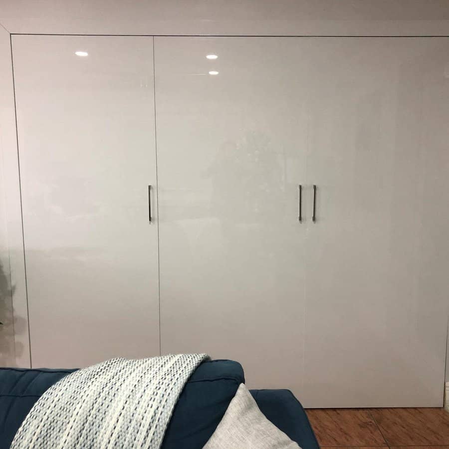 Sleek floor-to-ceiling white gloss closet doors with minimalist handles blend seamlessly into a modern living space with a cozy blue sofa