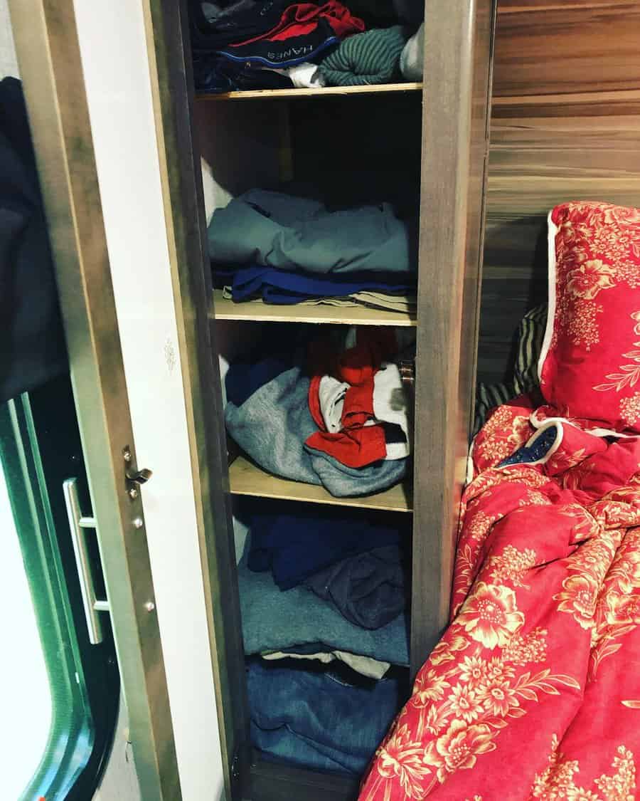 Small RV closet with open shelves storing folded clothes, located beside a bed with red floral bedding, optimizing vertical space.