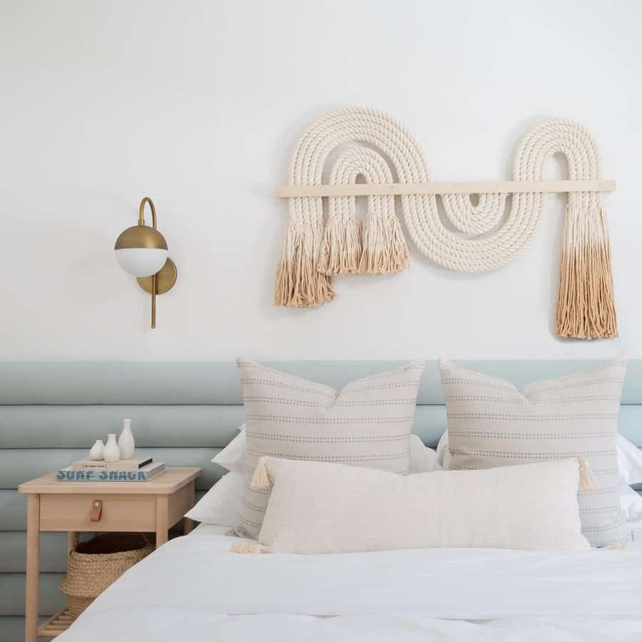 Bedroom with a light blue cushioned headboard, white pillows, woven wall art, bedside table, and a brass wall sconce