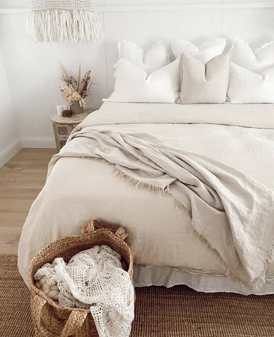 Minimalist bedroom with a cream bedspread, white pillows, woven basket with crochet items, and dried flowers on a bedside table