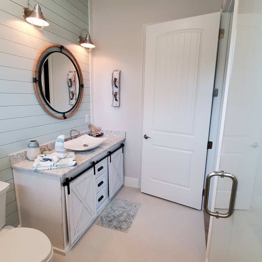 Bathroom with a round mirror, vanity with sink, light fixtures, and barn-style cabinet doors; shower with glass door on the right