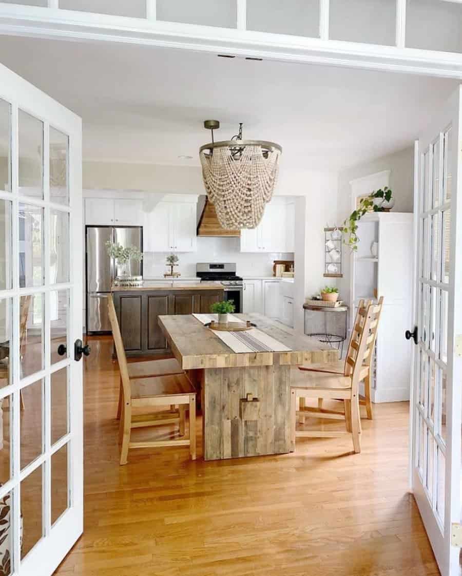 Bright kitchen and dining area with wooden table and chairs, beaded chandelier, and French doors leading in: white cabinets, plants, decor