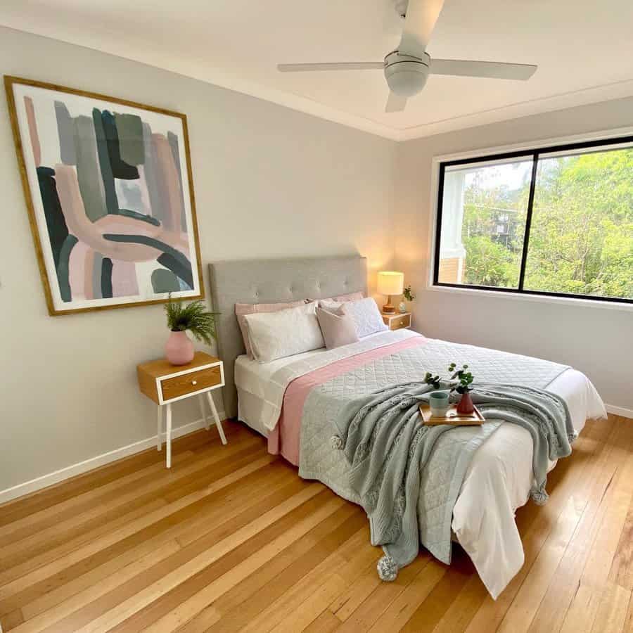 A cozy bedroom with a large window, double bed with pink and gray bedding, bedside table, abstract painting, and a ceiling fan