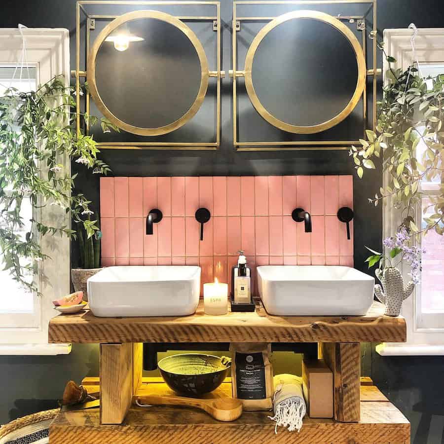 Modern double-sink bathroom with pink tile backsplash, black fixtures, gold mirrors, wood vanity, and lush greenery.