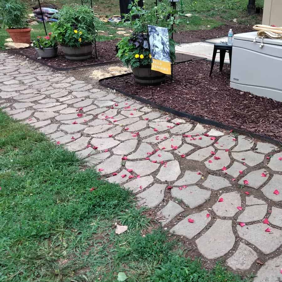 Stone garden path with scattered red petals surrounded by grass, potted flowers, and mulch