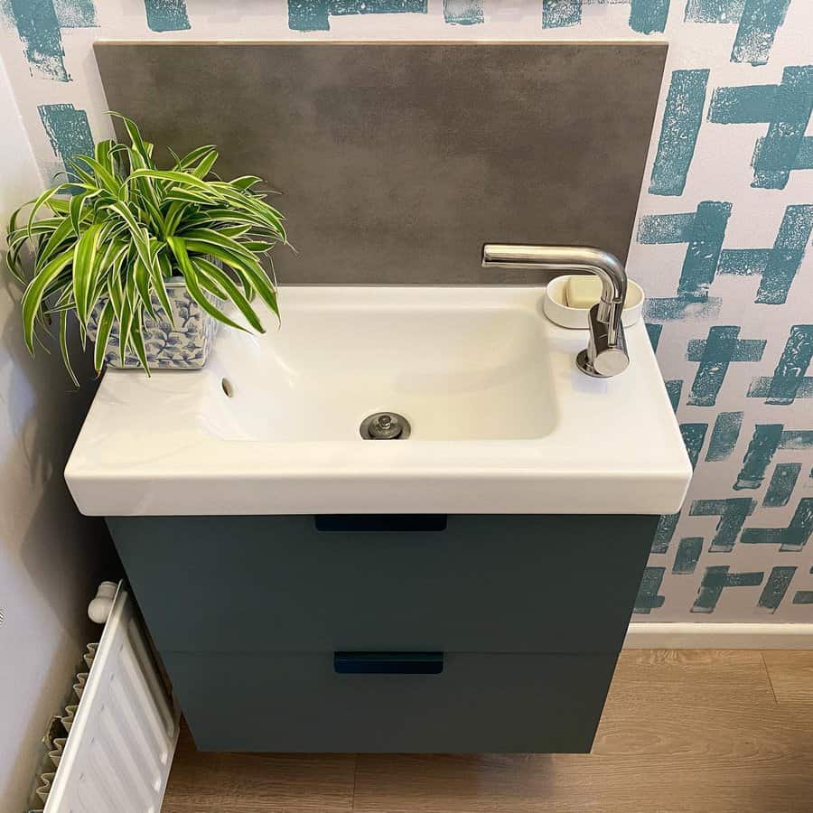 Compact bathroom sink with concrete backsplash, teal vanity, chrome faucet, and a spider plant adding a fresh, modern touch.