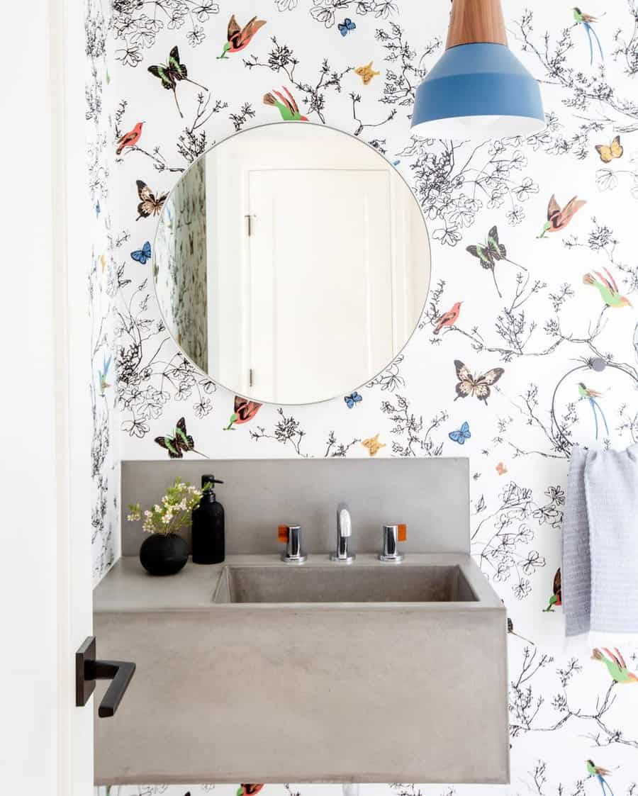 Whimsical bathroom with butterfly wallpaper, wall-mounted concrete sink and backsplash, round mirror, and modern blue pendant light.