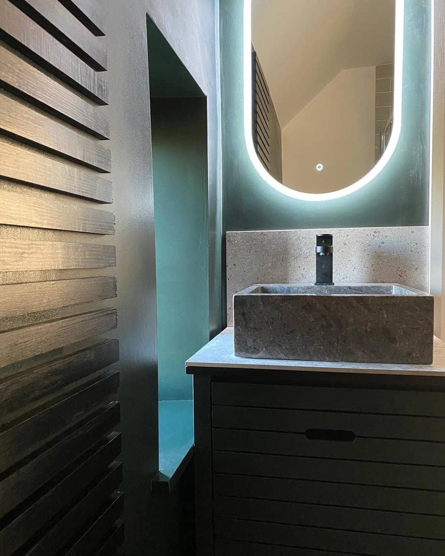 Moody modern bathroom with concrete backsplash, stone vessel sink, backlit oval mirror, and dark wood slatted vanity.