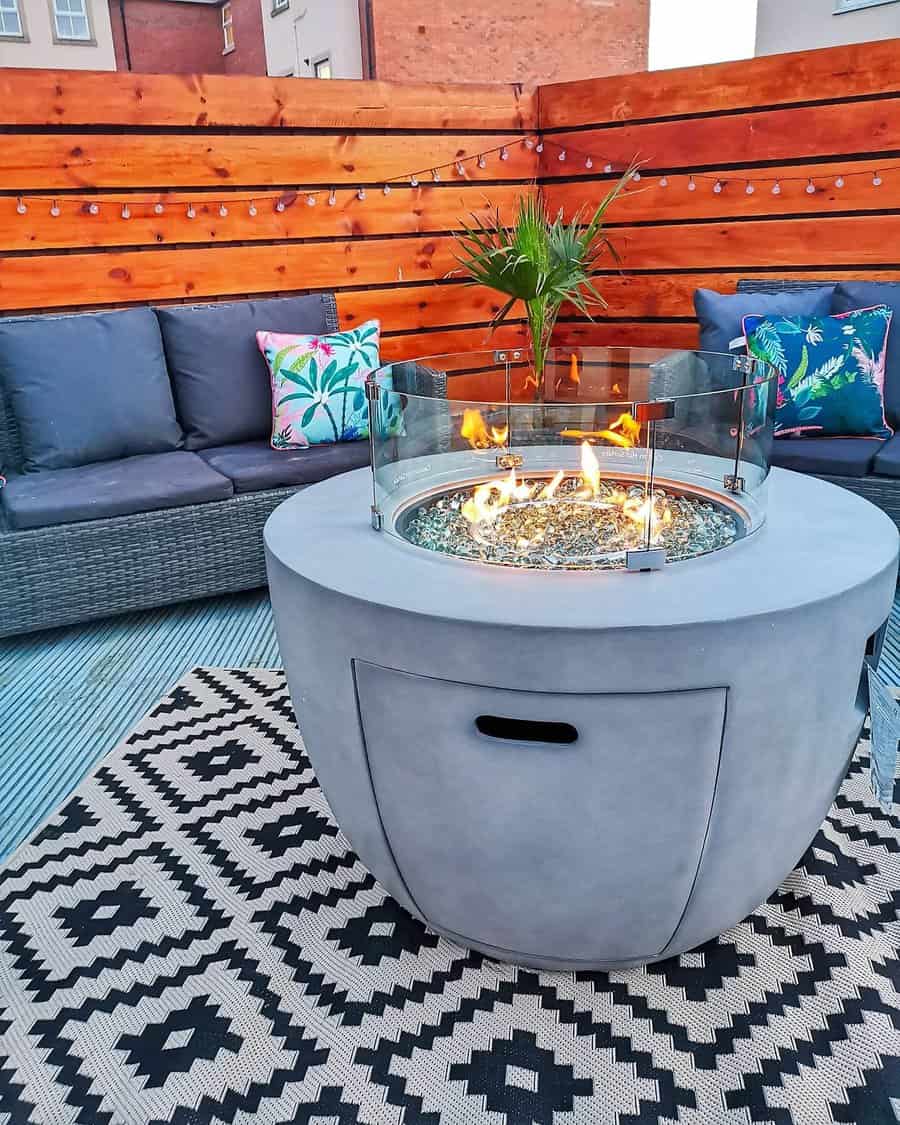 Rooftop patio with a round fire pit, cozy gray seating, colorful pillows, a patterned rug, and a wooden privacy fence