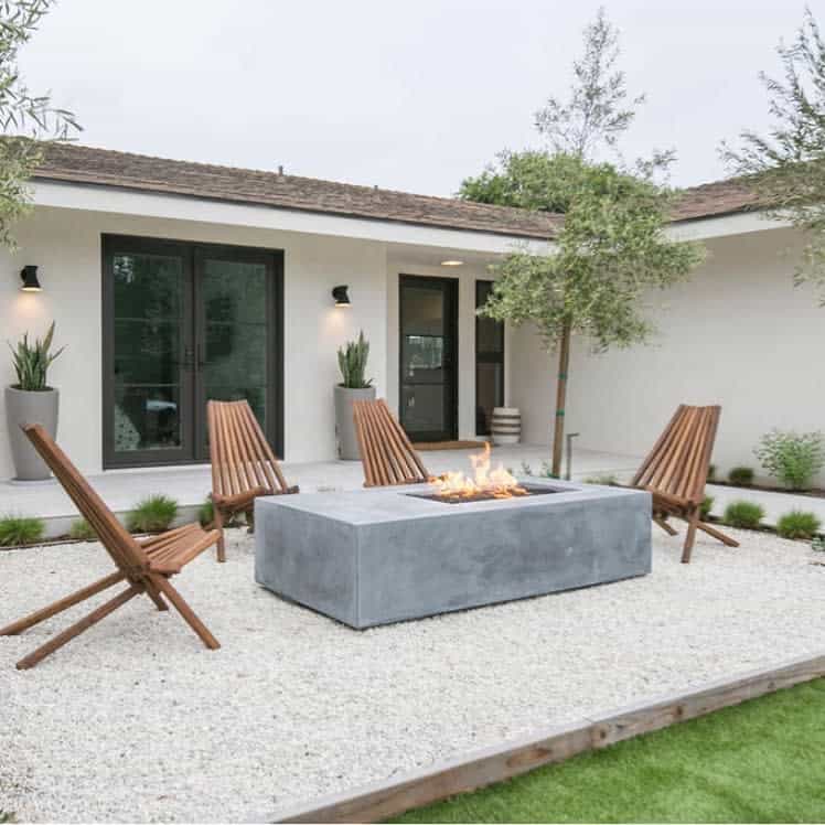 Outdoor patio with four wooden lounge chairs around a rectangular concrete fire pit, framed by greenery and a modern house facade