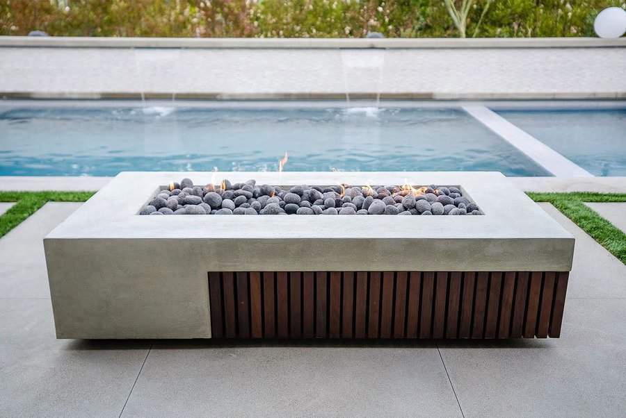 Modern rectangular fire pit with stones, set on a patio by a swimming pool with green hedges in the background