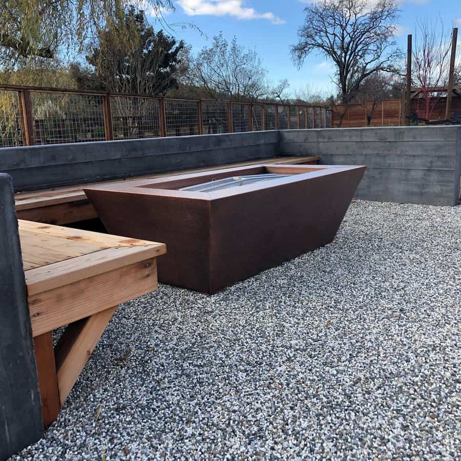 Modern outdoor fire pit with a sleek design, surrounded by wooden benches on a gravel surface, set against a backdrop of trees