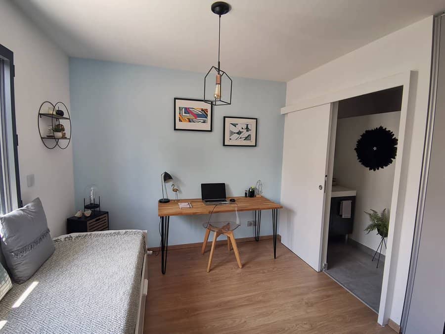 Home office with a wooden desk, laptop, and chair; minimalist decor includes framed art, a round wall shelf, and a pendant light