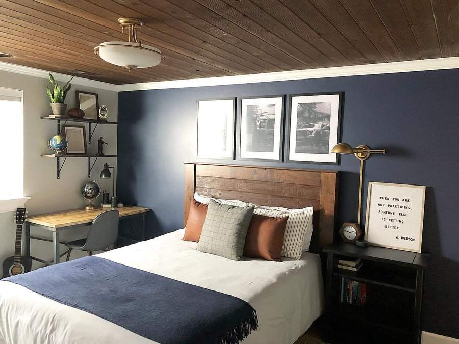 Cozy bedroom with a wood-paneled ceiling, a bed with blue and white bedding, a desk with a globe, and framed art on a dark wall