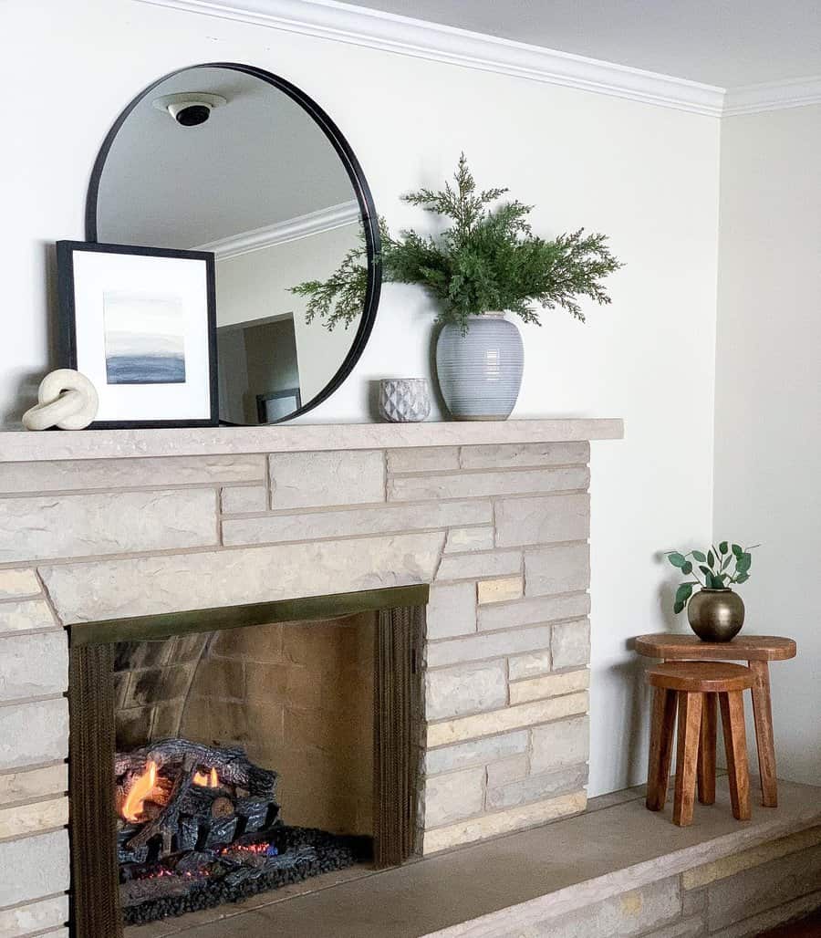 Cozy living room with a stone fireplace, round mirror, framed art, vase with green plant, small stool, and decorative items