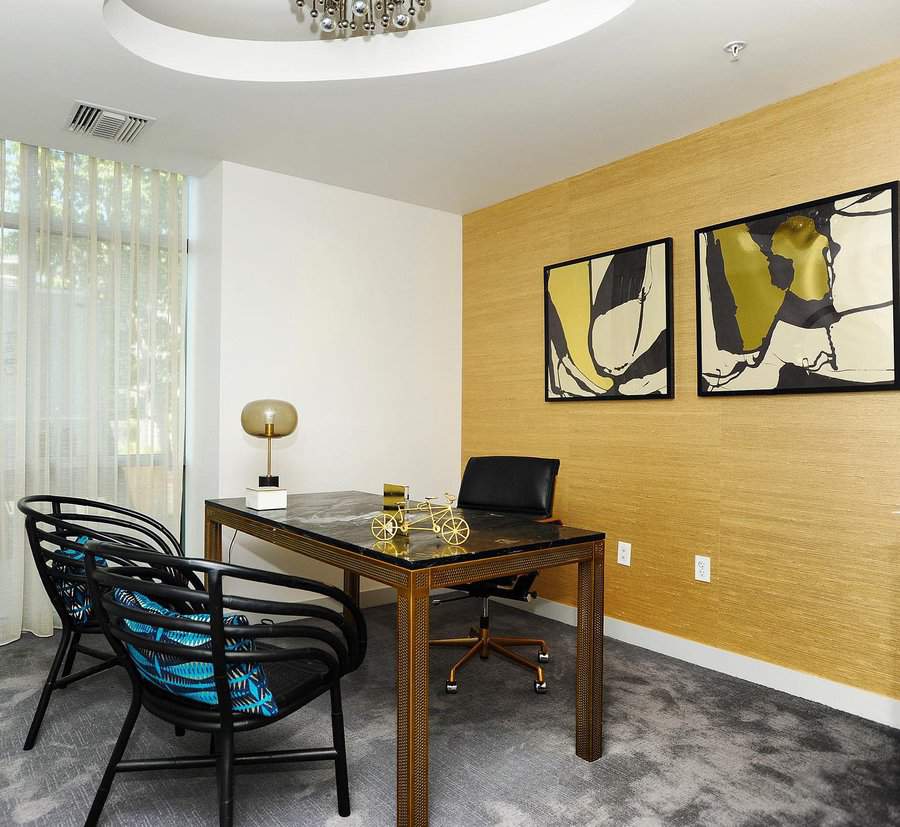 Modern office with a central desk, two black chairs, abstract art on the walls, and a lamp on the desk. Beige walls and curtain