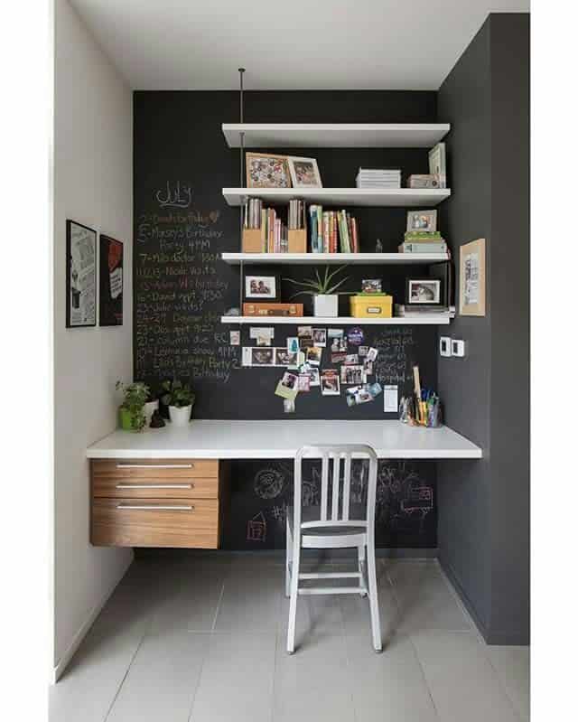 Modern home office nook with a white desk, wooden drawers, metal chair, black chalkboard wall, and three white shelves with books and photos