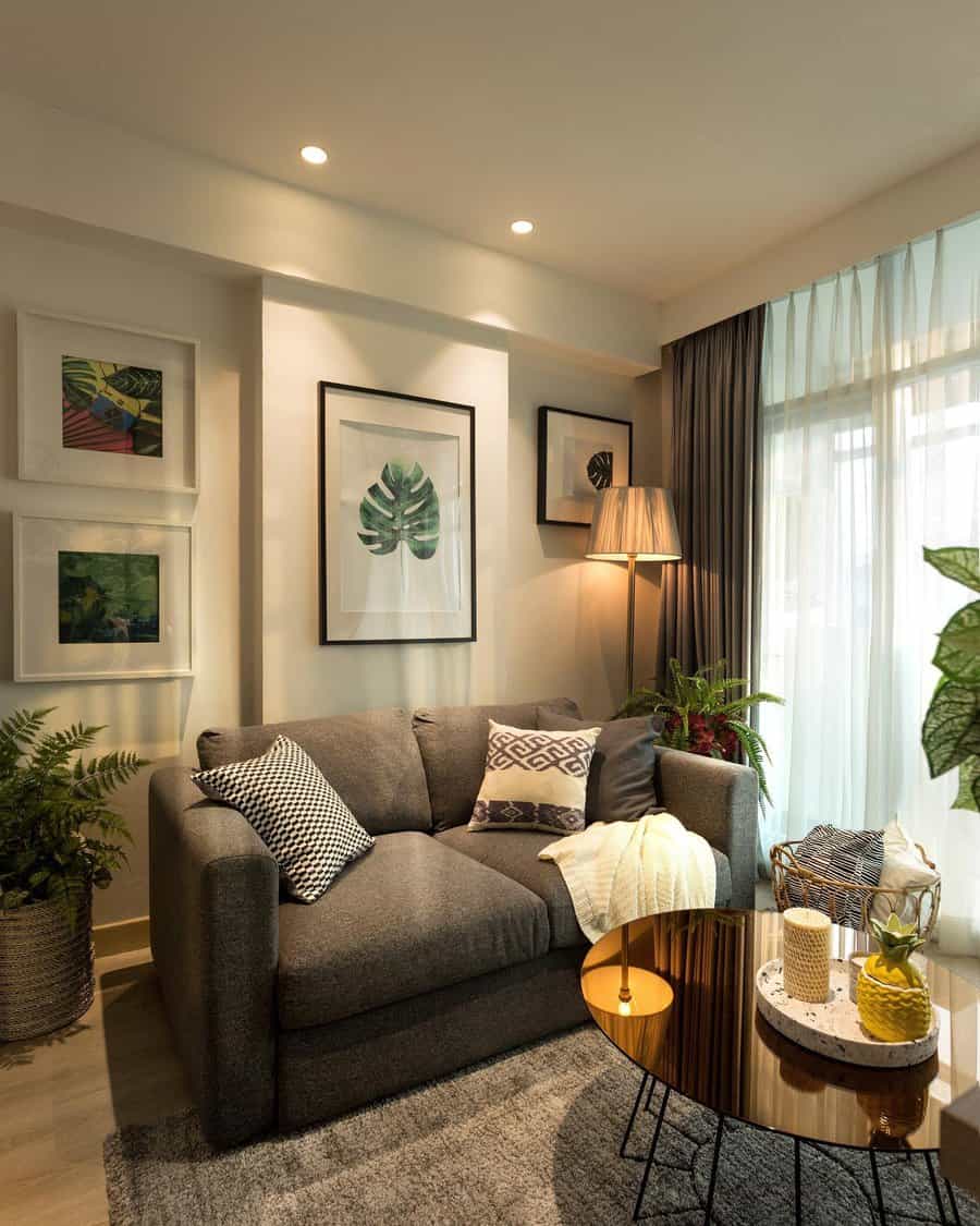 Small living room with a gray sofa, patterned pillows, plants, framed art, round coffee table, and floor lamp by a window