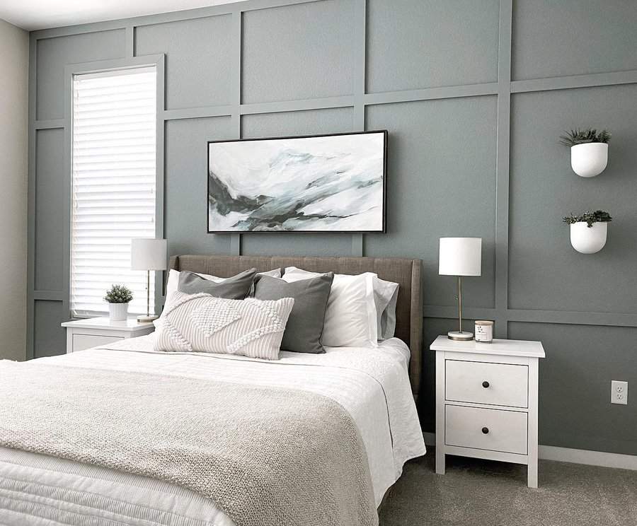 Minimalist bedroom with gray paneled wall, abstract art, white nightstands, lamps, and a bed with white linens and gray pillows