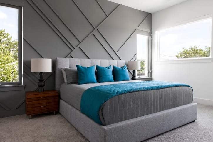 Modern bedroom with geometric accent wall, gray bed, blue bedding, and two bedside lamps; large windows with natural light