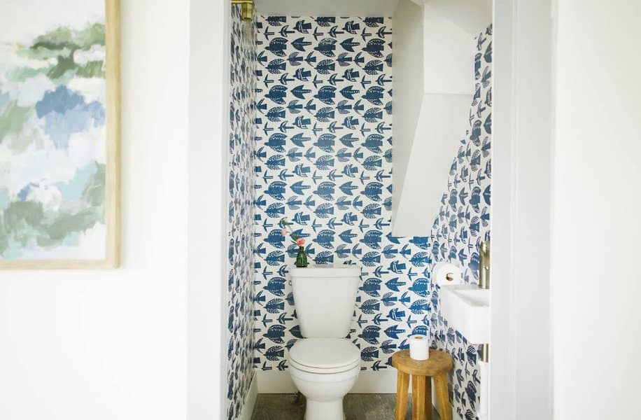 Small bathroom with bird-patterned wallpaper, white toilet with a flower vase on top, and a wooden stool by the sink