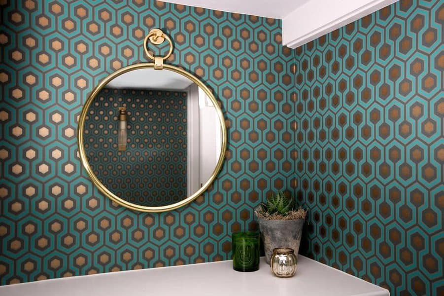 Round mirror on a wall with teal and gold geometric wallpaper, reflecting part of the room. Small plants and candles on a white shelf