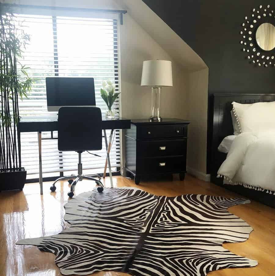 Bedroom with a zebra-print rug, desk, chair, lamp, and bed; bright window with blinds and bamboo plant in the corner