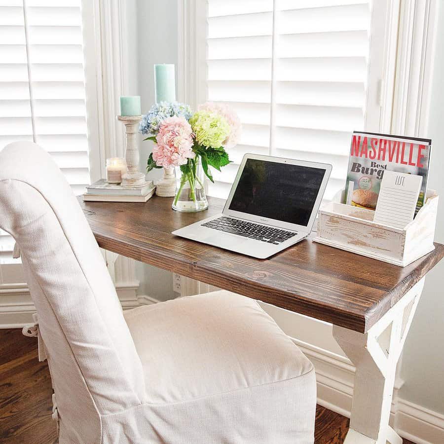 Cozy workspace with a laptop, flowers, and magazines on a wooden desk; cream chair and shutters in the background