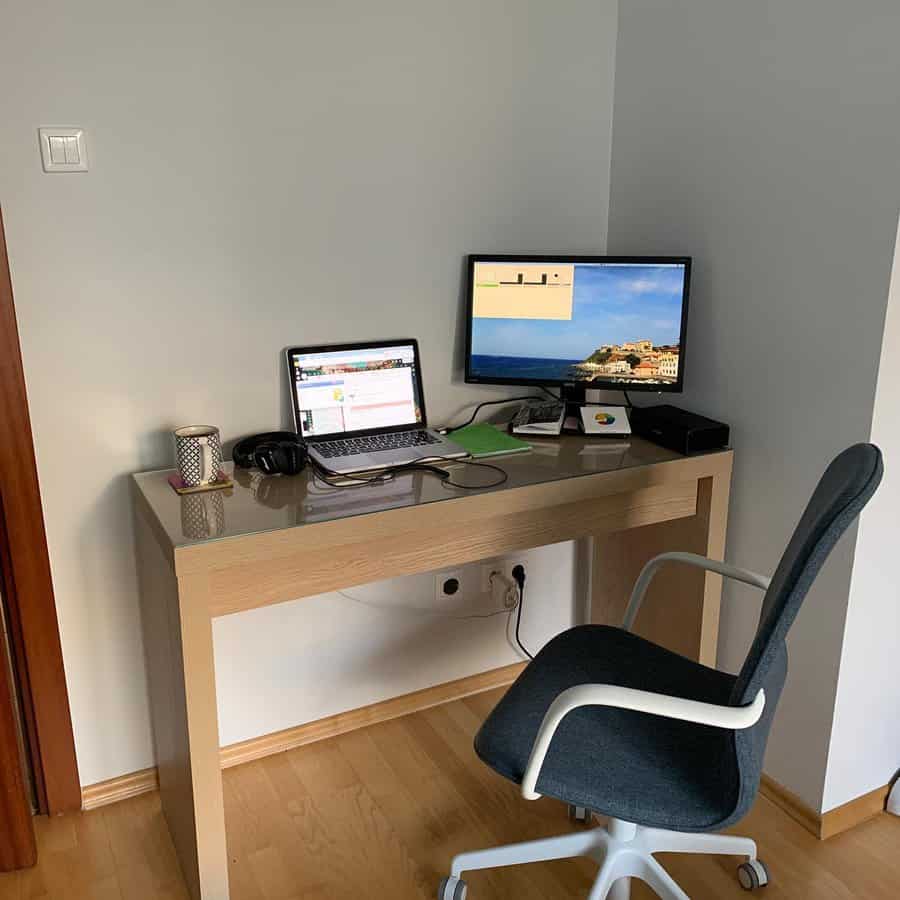 A tidy home office corner with a wooden desk, chair, laptop, monitor, headphones, mug, and a scenic wallpaper on the screen