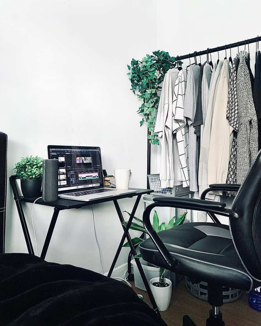 Home office setup with a laptop on a small desk, surrounded by plants and clothing on a rack, next to a black office chair