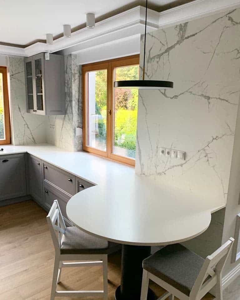 Elegant corner kitchen with a white round breakfast bar, marble walls, grey cabinetry, and large windows bringing in natural light