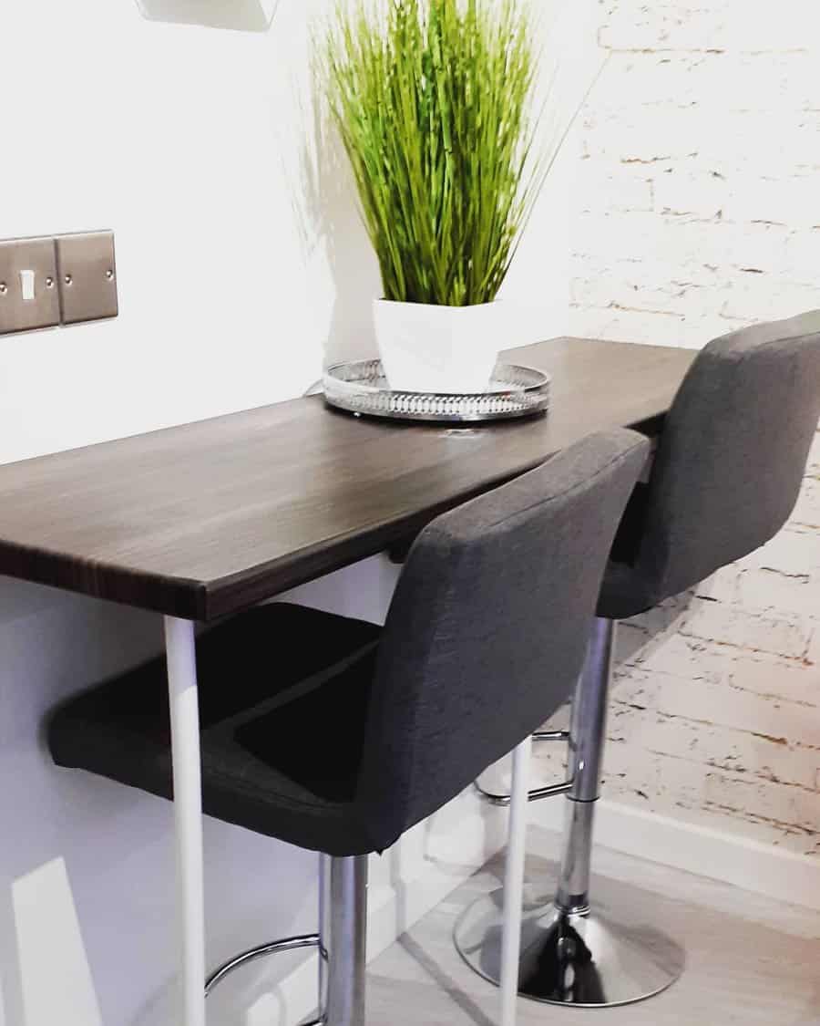 Two gray bar stools at a dark wooden counter, with a potted plant on a tray, against a white brick wall